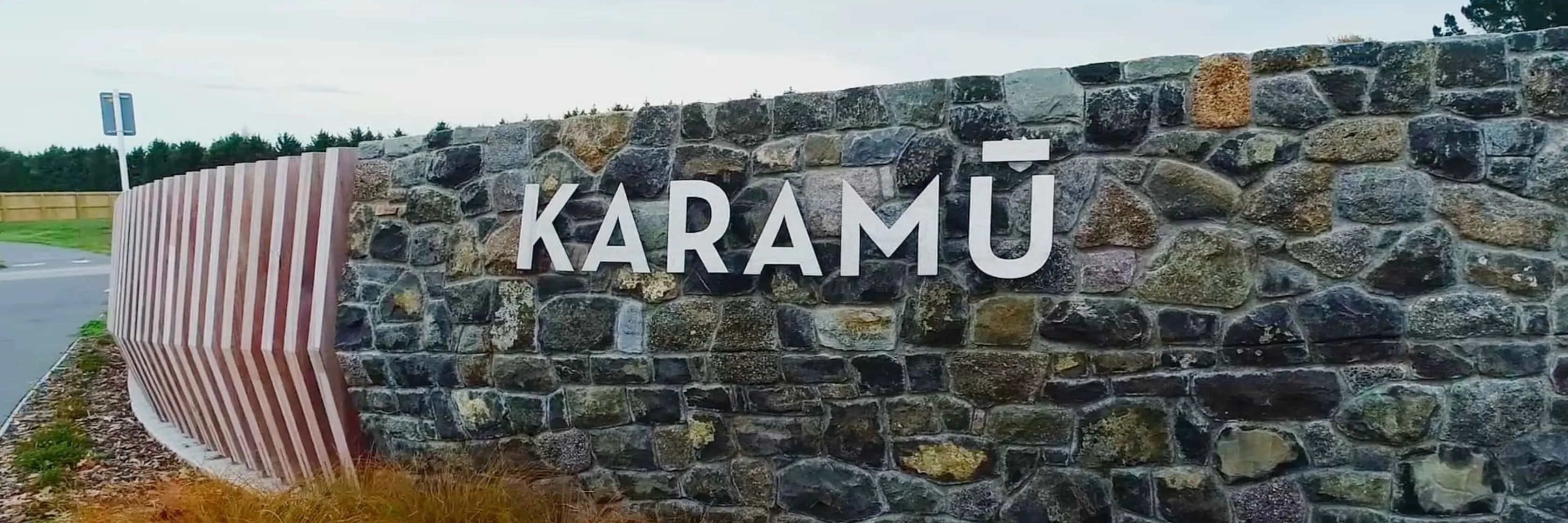 A stone wall with the word "KARAMŪ" in large white letters, located at the side of a road. Grassy vegetation is visible at the base of the sign. This geotechnical engineering project showcases work done by Eliot Sinclair's team of skilled geotechnical engineers.