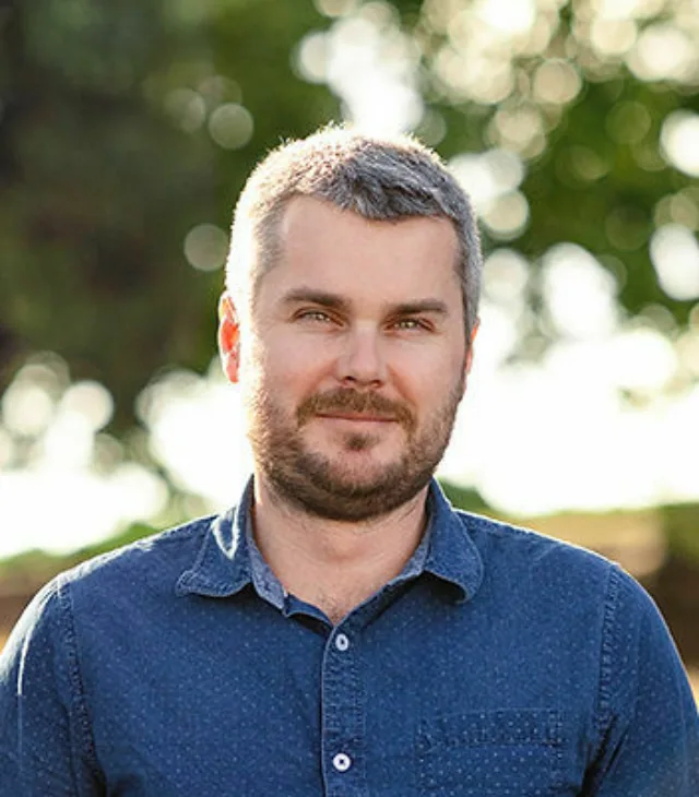 Ryan Orange, a Civil & Geotechnical Technologist at Eliot Sinclair, stands outdoors in a blue button-up shirt. His short hair and beard give him a rugged look, while trees blur softly in the background.