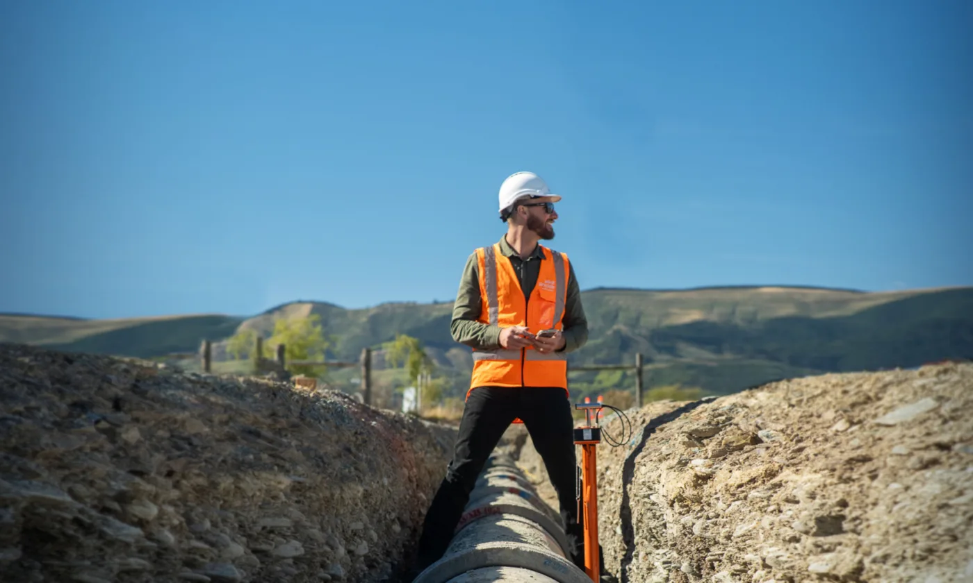 A geotechnical engineer wearing a white hard hat and an orange safety vest stands on a construction site trench with pipes, against a backdrop of Christchurch's hills and clear blue sky.