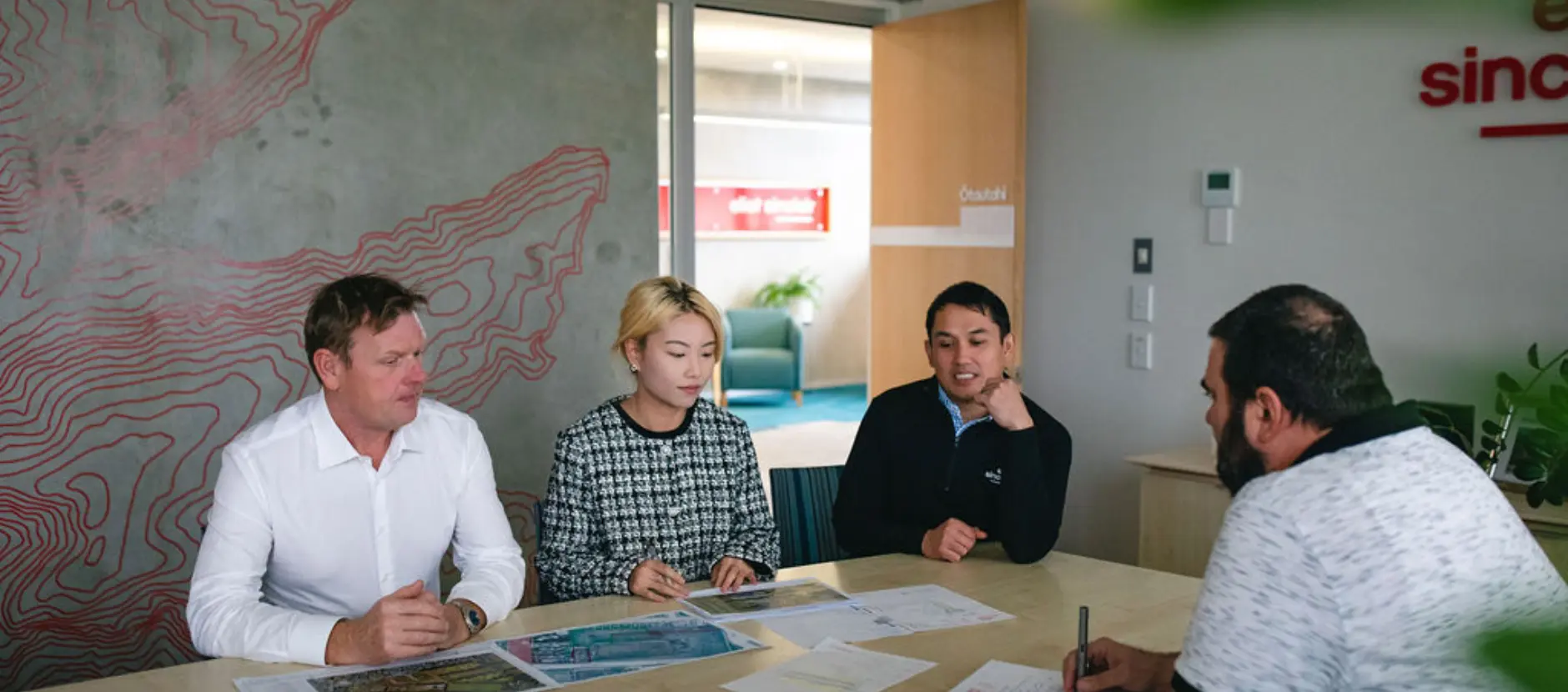 Four geotechnical engineers from Eliot Sinclair, sit around a table in a modern office, engaged in a meeting with various documents spread out before them. One person takes notes while the others discuss.