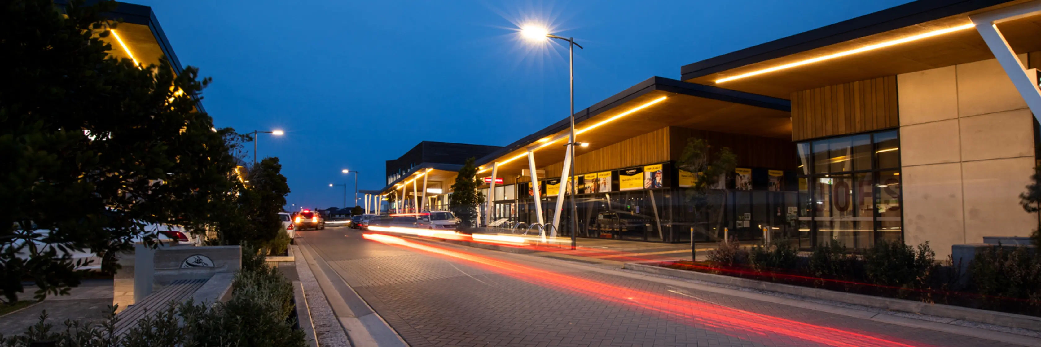 A photograph of Wigram Skies at dusk with trailing car lights, modern buildings on either side, and a clear, deep blue sky overhead. Streetlights illuminate the scene.