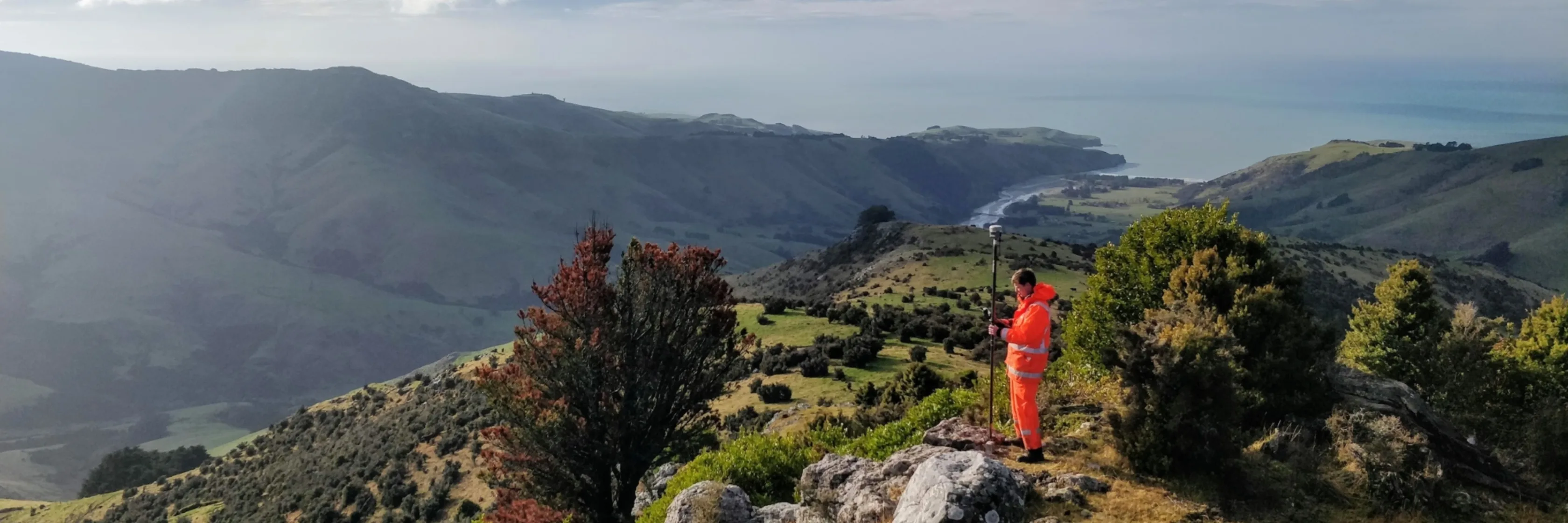 An Eliot Sinclair Land Surveyor Engineer in orange hi-vis clothing stands on a rocky hill, surrounded by green hills and valleys in Okains Bay, NZ, overlooking a distant body of water under a partly cloudy sky, capturing the breathtaking view.